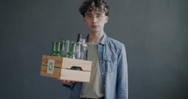 Cam şişelerle dolu bir kutu tutan genç adamın yavaş çekimde portresi ve gri arka planda Save the Planet tabelasını kaldırması. Ekoloji ve aktivizm kavramı.