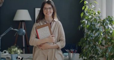 Evde elinde çizim kitaplarıyla kameraya bakıp gülümseyen neşeli genç kadın ressamın yavaş çekim portresi. Yaratıcı gençlik ve duygu kavramı.