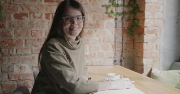 ノートと鉛筆の笑顔でカフェに座っている美しい若い女性のスローモーションポートレートカメラを見て カフェテリアでリモートワークを楽しむアーティスト — ストック動画