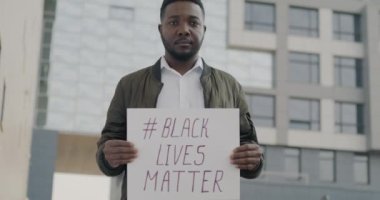 Siyahların Yaşamları Önemli Afro-Amerikan bayrağını taşıyan ciddi Afro-Amerikan bir adamın yavaş çekimde çekilmiş portresi kentsel arka planda duruyor. Irkçılık ve eşitlik kavramı.