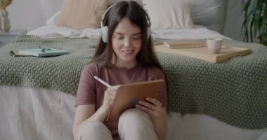 Grafik tabletinde çalışan ve yatak odasında kulaklıkla müzik dinleyen serbest çalışan bir kadın. Yaratıcı meslek ve serbest çalışma kavramı.