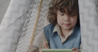 Akıllı telefonlu video oyunu oynayan sevimli bir çocuk ve evde çocuklar için komik bir çadırda oturmuş gülümsüyor. Malzemeler ve eğlence konsepti.