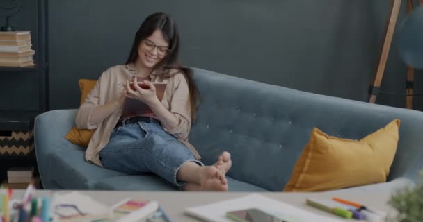 アパートのソファでゆったりとした時間を過ごすことができるタブレットを使って 幸せな若い女性デジタルアーティスト 家庭生活と近代技術の概念 — ストック動画