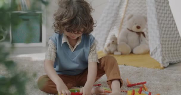カーペットに座って家の中で木製のおもちゃで遊んでいる巻き毛のある愛らしい子供 子どもと保育園のインテリアコンセプトの開発 — ストック動画