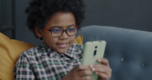 スマートフォンのタッチスクリーンを使用してかわいいアフリカ系アメリカ人の子供は 自宅でソファに座って笑顔 現代のガジェットや子供時代のコンセプト — ストック動画