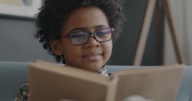 Gözlüklü, kitap okuyan ve salonda gülümseyen neşeli Afrikalı Amerikalı çocuk. Edebiyat ve zeki çocuk konsepti.
