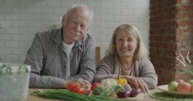 Mutfak masasında oturan ve kameraya gülümseyen güzel yaşlı çiftin yavaş çekimde portresi. Emeklilik ve sağlıklı beslenme konsepti.