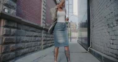 Moda kıyafetli, açık havada dans eden ve kulaklıkla müzik dinleyen neşeli bir kadının yavaş çekim portresi. Eğlence ve modern gençlik kavramı.