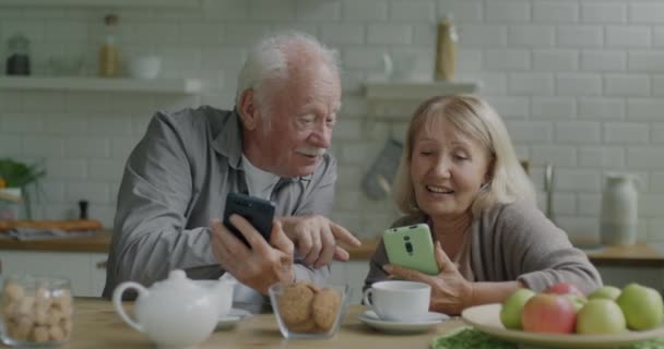 スマートフォンを使ってディスプレイを見せ キッチンテーブルに座って話をするシニア男性と女性 近代的な技術と退職の概念 — ストック動画