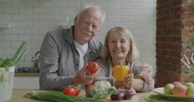 Mutfak masasında birlikte oturan, elinde taze sebzelerle gülümseyen emekli bir adam ve kadının yavaş çekimde portresi. Emeklilik ve beslenme kavramı.