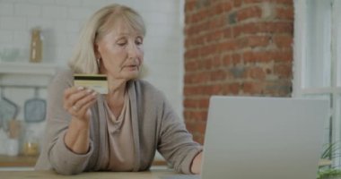 Yaşlı bir kadın, evin mutfağında dizüstü bilgisayarla alışveriş yaparak kredi kartıyla ödeme yapıyor. Emeklilik ve bankacılık kavramı.