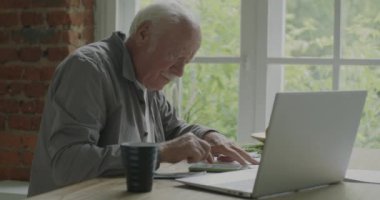 Ev taksitlerini sayan yaşlı bir adam akıllı telefon kullanarak evde oturmuş paraları tutan faturalara bakıyor. Aile bütçesi ve finans kavramı.