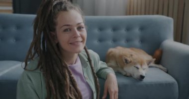 Arka planda kanepede Shiba inu köpeği yatarken rastalı saçlarıyla gülümseyen güzel genç bir kadının yavaş çekimde portresi. İnsanlar ve evcil hayvanlar kavramı.