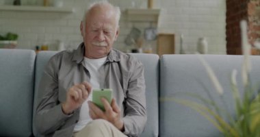 Modern apartmandaki koltukta oturan akıllı telefondan mesaj atan yaşlı bir adam. Çevrimiçi içerik ve internet iletişim kavramı.