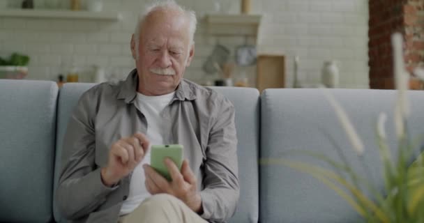 在现代公寓里 老年人坐在沙发上 用智能手机触摸屏幕发短信 在线内容和互联网通信概念 — 图库视频影像