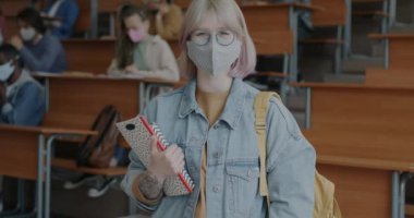 Kolejde elinde kitap tutan, tıp maskesi takan kadın öğrencinin yavaş çekimde portresi. Halk ve pandemik kısıtlamalar kavramı.