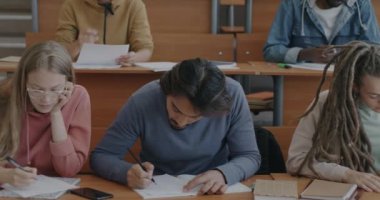 Çoklu etnik gruptan erkek ve kadın öğrenci sınav yazma testleri konferans salonunda masada oturuyorlar. Akademik eğitim ve öğrenim kavramı.