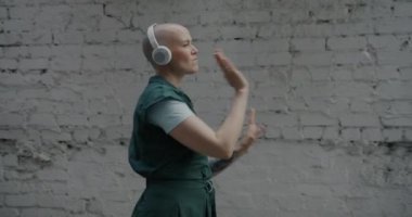 Kulaklıklı şık bir kadının yürümesi ve dans etmesi ve tuğla duvarın yanında müzik dinlemesi. Gençlik ve kentsel yaşam tarzı kavramı.