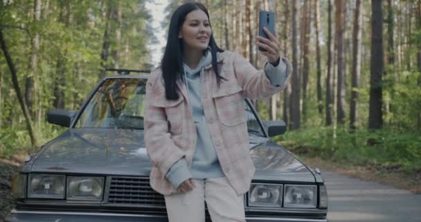 快乐的女孩在网上用智能手机打电话 谈论公路旅行 站在森林路上的汽车旁边 — 图库视频影像