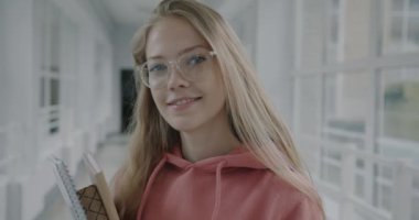 Lise koridorunda elinde kameraya gülümseyen kitaplarla duran gözlüklü genç bir kadının portresi. Halk ve eğitim kavramı.