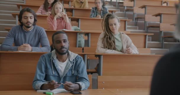 他の学生と一緒に大学のホールに座って教師に話す質問をするアフリカ系アメリカ人の陽気な男 専門的な議論と教育の概念 — ストック動画