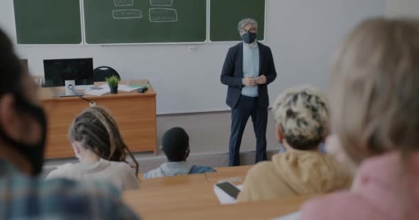 コロナウイルスのパンデミックの間に教室で学生の顔マスク指導グループを身に着けている英語の教師成熟した男 人々と教育の概念 — ストック動画