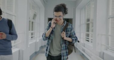 Üniversite koridorunda yürüyen, cep telefonuyla eğitim hakkında konuşan neşeli bir gencin yavaş çekim portresi. İnsanlar ve iletişim konsepti.