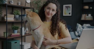 Evdeki masada oturan ve elinde sevimli Shiba Inu köpeğini tutan güzel bir kadının yavaş çekimde portresi. Evcil hayvanlar ve insanlar konsepti.