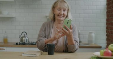 Akıllı telefon ekranı araklayan ve mutfakta çay içen son sınıf kadınının portresi. Modern aletler ve emekli insanlar konsepti.