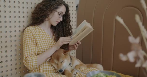 若い女性が本を読んで ペットフレンドリーなカフェでテーブルに座って柴犬を撫でる レジャー活動と動物コンパニオンコンセプト — ストック動画