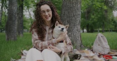 Yavaş çekimde gülümseyen genç bir kadının portresi sonra da yeşil parkta sevimli Shiba Inu köpeğini öpüyor. Hayvanlar ve sevecen insanlar kavramı.