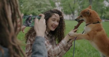 Kız kardeşinin yeşil parkta Shiba Inu köpeğiyle oynarken fotoğrafını çekiyor. Akıllı telefon kamerasıyla ekrana dokunuyor. Teknoloji ve fotoğraf konsepti.