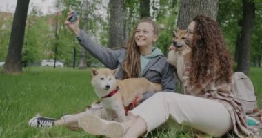 Genç bayan arkadaşlar Shiba Inu köpekleriyle selfie çekiyorlar akıllı telefon kameralarına poz veriyorlar parkta evcil hayvanlarla eğleniyorlar. Arkadaşlık ve hayvanlar konsepti.