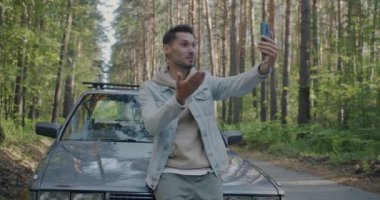 Mutlu genç adam el sallayarak online video görüşmesi yapıyor. Akıllı telefonuyla dışarıda, ormanda, otomobilin yanında. Seyahat ve iletişim konsepti.