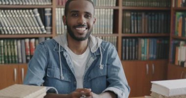 Modern kütüphanede oturmuş kameraya gülümseyen neşeli Afrikalı Amerikalı mutlu öğrencinin yavaş çekim portresi. Halk ve eğitim kavramı.