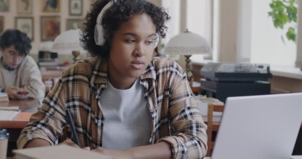 アフリカ系アメリカ人の女子学生がノートにノートを書き 図書館のヘッドフォンで音楽を聴く 現代のライフスタイルと教育の概念 — ストック動画