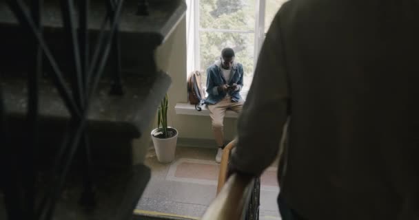 男はまだガジェットを使用して窓の上に座っている間 男性学生は大学ホールで階下を歩く 人とキャンパスライフスタイルのコンセプト — ストック動画