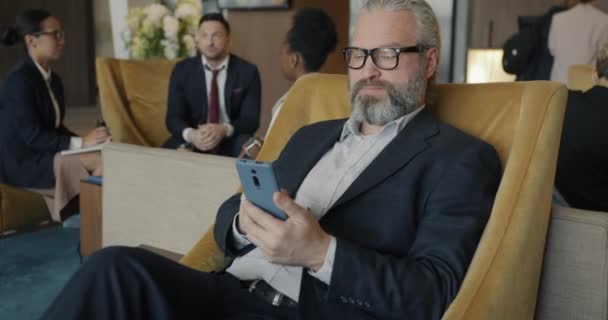 成功的商人正在酒店大堂与一群穿着西装的人一起使用智能手机发短信放松心情 现代技术和商人概念 — 图库视频影像