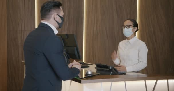医療マスクを身に着けているビジネスマンは パンデミックの間 フロントのホテルマネージャーと話しています 宿泊施設とコロナウイルスの概念 — ストック動画
