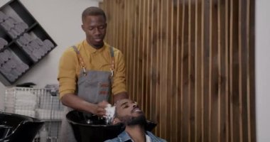 Berber dükkanında müşteriyle ilgilenmek için şampuanla saç yıkayan yetenekli bir berber. Afro-Amerikalı adam modern salonda tedavi görüyor..