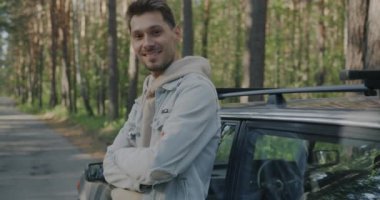 Ormanda modern otomobilin yanında durup kameraya bakıp gülümseyen neşeli genç bir adamın ağır çekimde portresi.