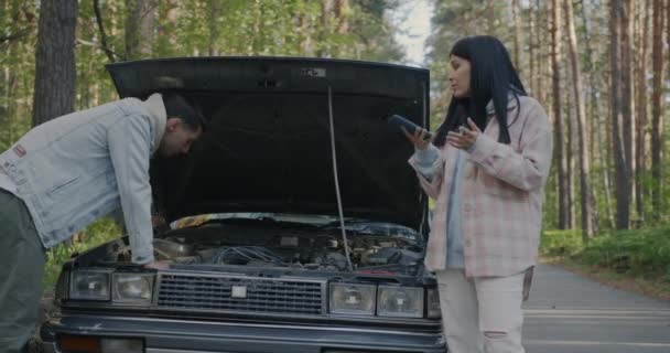 男人试图修理破碎的汽车 而女人在外面的森林路上用手机求助 运输和事故概念 — 图库视频影像