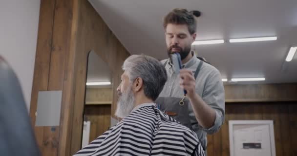 愉快的理发师在理发店里用吹风机和刷子与客户一起工作 微笑交谈 优雅的绅士得到专业的待遇 — 图库视频影像