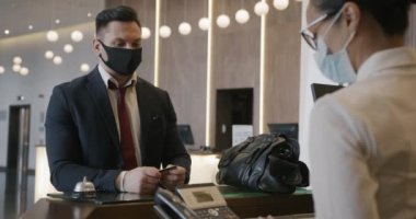 Tıbbi maske takan bir otel müşterisi resepsiyonistle konuşuyor sonra da kredi kartıyla ödeme yapıyor ve gidiyor. İnsanlar ve salgın konsepti.