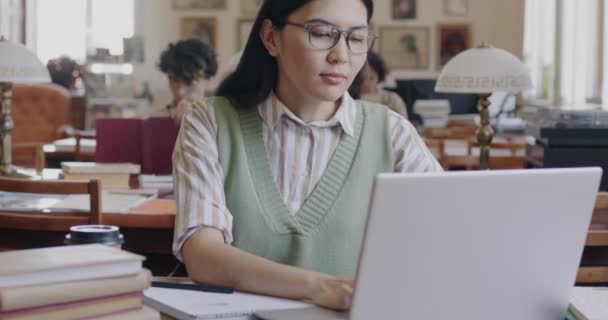 アジアの女子学生は ノートパソコンのタイピングや大学図書館でのスマートフォンの勉強をしています 現代の装置と学術教育の概念 — ストック動画