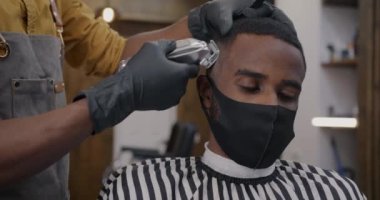 Tıbbi maske takan genç bir adam saçını makineyle tıraş ettirirken berber de kuaförde çalışıyor. Salgın ve kısıtlamalar kavramı.