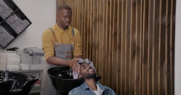 男性の美容師は ヘアカットの準備快適な理髪店でアフリカ系アメリカ人のクライアントに頭を乾燥させます 幸福と衛生の日常概念 — ストック動画