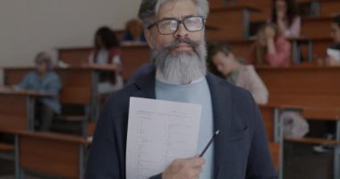 Kolejde öğrenciler sınav yazarken sınıfta elinde kağıtlarla duran olgun bir öğretmenin yavaş çekimde portresi. Eğitim ve sınav kavramı.