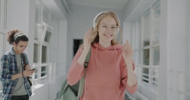 大学の廊下を歩いて踊るヘッドフォンを介して音楽を聞いて屈託のない若い女性のスローモーション 現代のライフスタイルと若者文化の概念 — ストック動画