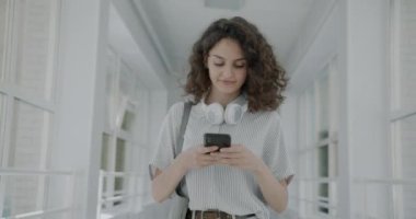 Dolly 'nin üniversite koridorunda akıllı telefon mesajlarıyla yürüyen mutlu genç kız fotoğrafı. Sosyal medya ve modern teknoloji kavramı.